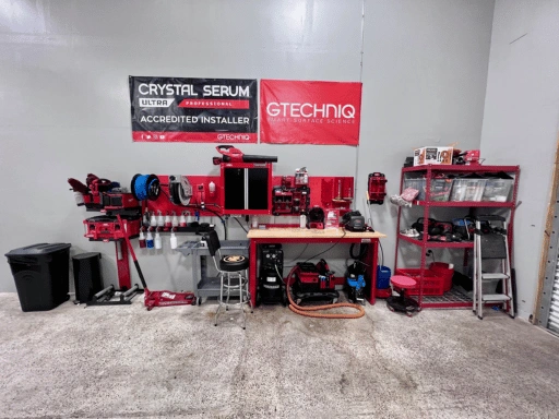 Close-up of Vibrant Mobile Detail ceramic coating workstation showing GTechniq Crystal Serum Ultra Professional Accredited Installer banner and GTechniq Smart Surface Science banner on the wall, red pegboard tool organizer, polishing machines, hose reel, workbench with bottles, and red supply shelving.