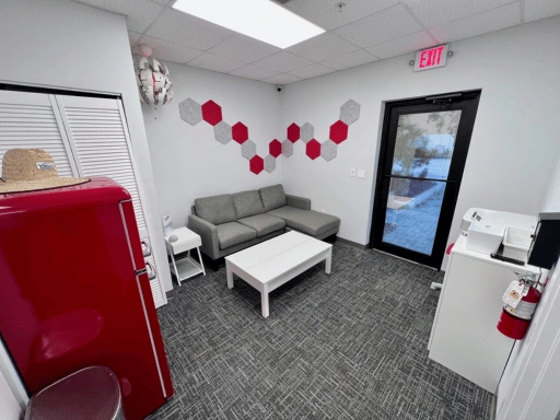 Another angle of Vibrant Mobile Detail customer lounge showing the front entrance glass door, gray sofa with white coffee table, red retro refrigerator, decorative hexagon wall art in red and gray, pendant light, and printer station area.