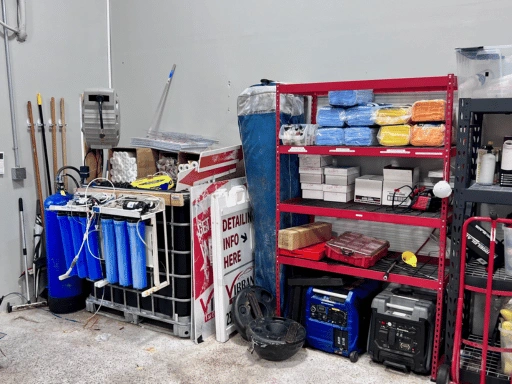 Vibrant Mobile Detail equipment corner showing blue multi-stage deionized water filtration tanks, portable generators, a red metal shelving unit with stacked microfiber towels and boxed supplies, extension poles, and a Vibrant branded sign.