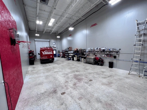 Wide alternate angle of Vibrant Mobile Detail shop bay showing the branded red Ford Transit van near the roll-up door, red accent wall with mounted basketball hoop, full equipment and supply wall with workbench and shelving, extension ladder, and high ceiling with industrial lighting.