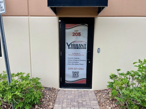 Exterior entrance to Vibrant Mobile Detail shop at Unit 205 showing a branded glass door with the Vibrant logo, service list including ceramic coating interior and exterior detailing paint correction and fleet services, phone number 239-221-6364, website URL, QR code, surrounded by tropical landscaping.