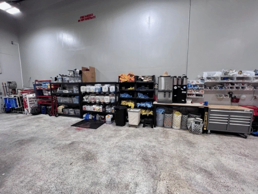 Organized supply wall inside Vibrant Mobile Detail shop showing metal shelving units stocked with detailing chemicals, color-coded microfiber towels in blue orange and yellow, storage bins, a professional Husky tool chest, and workbench area.