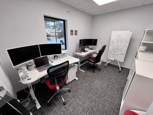 Vibrant Mobile Detail office with two workstation desks featuring dual and triple monitor setups, ergonomic chairs with red accents, a whiteboard with business planning notes, and organized shelving in a carpeted office space.