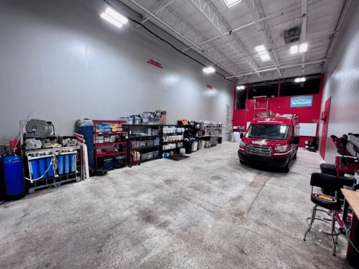 Wide view of Vibrant Mobile Detail’s Fort Myers detailing shop showing a branded red Ford Transit van parked inside, organized equipment shelving along the wall, blue deionized water filtration tanks, and overhead industrial lighting in a spacious air-conditioned facility.