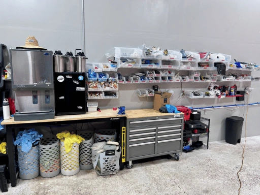 Close-up of Vibrant Mobile Detail workbench showing wall-mounted clear storage bins with small parts and accessories, a Husky tool chest, water dispensers, butcher block countertop, and laundry baskets filled with sorted microfiber towels.